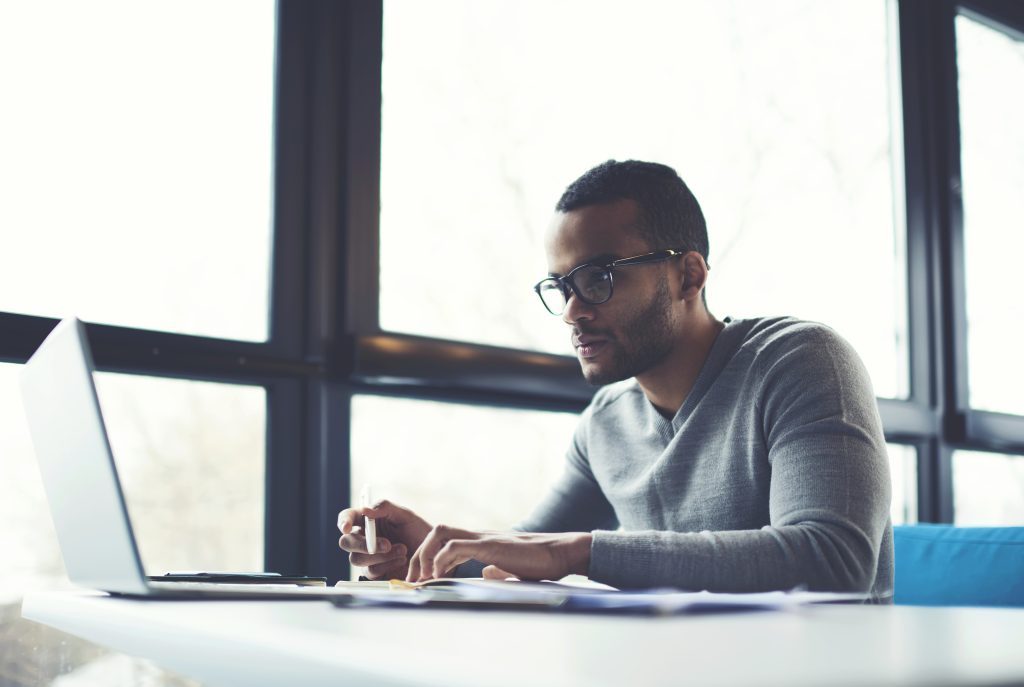 Concentrated businessman checking accounting documentation in online database on modern computer connecting to wireless internet connection. Male aro american entrepreneur working on laptop indoors